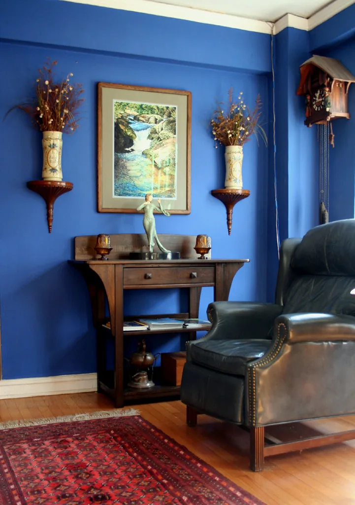 A blue reading room with an armchair, small table and framed painting.